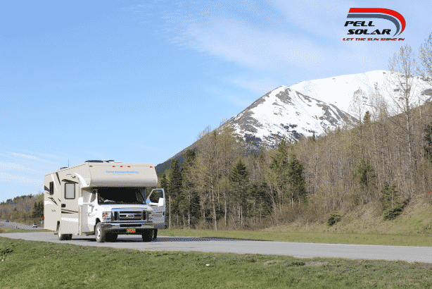 A tan RV is traveling down an open road with green grass and small forest trees beside the road. It is a sunny day, and a big mountain with snow on top of it is in the background.